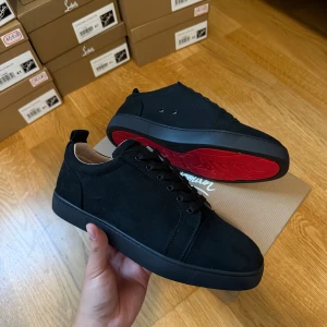 Svarta sneakers från Louboutin mocka storlek 45 - Snygga svarta sneakers från Christian Louboutin i mocka med klassisk röd sula och svarta snören. Skorna har en stilren design med rund tå och platt sula, perfekt för dig som vill sticka ut med exklusiva detaljer.