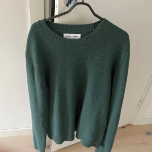 Grön stickad tröja från Samsøe Samsøe - Mysig mörkgrön stickad tröja från Samsøe Samsøe i storlek XL. Tröjan har rund halsringning, ribbade muddar och är tillverkad av återvunnen manchester. Perfekt för lager-på-lager och chill dagar.