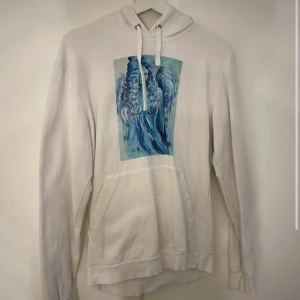 The cool elepante  - Vit hoodie med stor ficka framtill och huva med snörning. På bröstet finns ett blått konstnärligt tryck som ger en unik vibe och vill sticka ut lite extra.