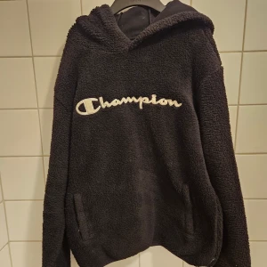 Ny oanvänd svart vintage fleece hoodie från Champion - Mysig svart hoodie från Champion i fluffig fleece med broderad logga framtill. Tröjan har huva, långa ärmar och två fickor på sidorna. Perfekt för kalla dagar när du vill vara både bekväm och snygg. Den har en vintage vibe och köptes för 1000 kr. Pris går att diskutera. Skriv ifall frågor uppkommer eller fler bilder behövs. Den är såklart äkta. 