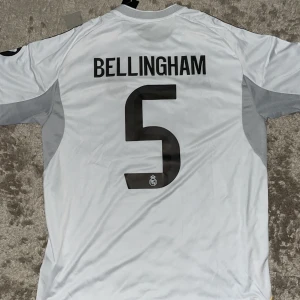 Real Madrid Bellingham tröja Adidas L - Säljer en officiell Real Madrid fotbollströja från Adidas med Bellingham och nummer 5 på ryggen. Tröjan är vit med svarta detaljer, gula linjer och grå partier på sidorna. Klassisk rund hals, korta ärmar och tillverkad i lätt funktionsmaterial (AEROREADY).
