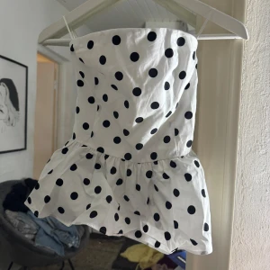 Vit prickig tubtopp från Bik Bok - Supersöt vit tubtopp från Bik Bok med svarta prickar och volang nertill. Aldrig använd och prislapp kvar. Toppen är bandeau-modell med smockad baksida för extra komfort och sitter snyggt på kroppen. Perfekt för dig som vill sticka ut med en lekfull och trendig look.