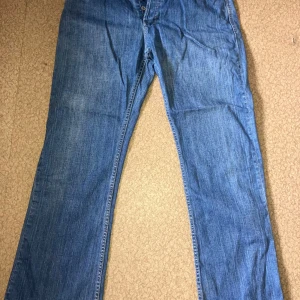 Blå jeans från Diesel, storlek 33 - Säljer ett par klassiska blå jeans från Diesel med snygga detaljer på bakfickorna och tydlig logga framtill. Jeansen har rak passform och är tillverkade i slitstarkt bomullsjeans. Perfekta för dig som gillar en tidlös denimlook. Fickorna har dock små hål