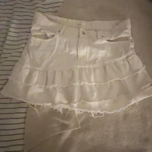 Vit volangkjol från Gina tricot - Supersöt vit kortkjol från Gina med volanger och rå kant nertill. Kjolen har klassiska jeansfickor och knapp framtill. Perfekt för dig som gillar en lekfull och trendig stil.