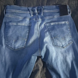 Replay Anbass blå jeans - Snygga blå Replay Anbass jeans med klassisk femficksdesign och ljusa slitningar. Jeansen har normal passform, raka ben och är tillverkade i mjukt denimtyg med snygga detaljer på bakfickorna. Perfekta för dig som gillar stilrena och bekväma jeans.