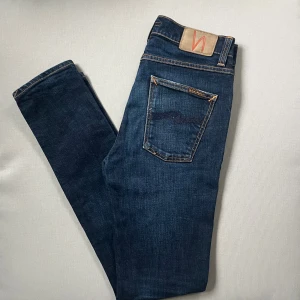 Nudie Jeans Lean Dean slim - Mörkblå jeans från Nudie Jeans, modell Lean Dean. Klassisk femficksdesign med orange kontrastsömmar och Nudie-logga på bakfickan. Jeansen har en slim passform och är tillverkade i mjukt denimtyg för en snygg och bekväm look. De har tecken på användning 
