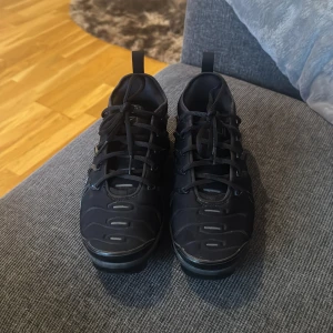 Nike Air Vapormax Plus Svarta Sneakers - Svarta Nike Air Vapormax Plus med futuristisk design och synlig luftdämpning i sulan. Skorna har snörning, diskreta loggor och en glansig overlay på ovandelen. Perfekta för dig som gillar streetwear och vill ha en riktigt clean look.