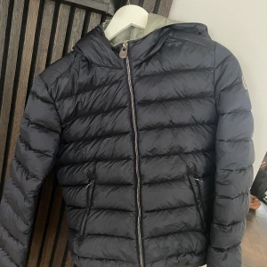 Marinblå colmar jacka - Marinblå dunjacka med huva och dragkedja framtill. Jackan har quiltad design med horisontella sömmar och två fickor med dragkedja. Perfekt för kyliga dagar och har en stilren look som funkar till det mesta. I bra skick förutom sista bilderna, inget som påverkar helhetsintrycket. Passar en S. Nypris 5000
