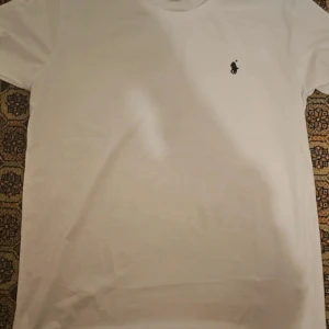 Vit t-shirt från U.S. Polo Assn. - Vit t-shirt från U.S. Polo Assn. i klassisk modell med rund hals och korta ärmar. T-shirten har en liten svart broderad logga på bröstet och är tillverkad i mjuk bomull. Perfekt basic-plagg med clean stil.
