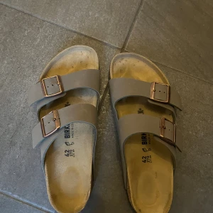 Birkenstock sandaler i beige, stl 42 - OANVÄNDA! Birkenstock sandaler i beige med två breda remmar och justerbara metallspännen i kopparfärg. Klassisk korksula och anatomisk fotbädd för extra komfort. Perfekta för sommaren och chill dagar. Snygg och enkel design med platt sula.