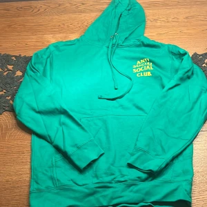 Grön hoodie från Anti Social Social Club - Säljer en äkta grön hoodie från Anti Social Social Club med stor gul logga på ryggen och mindre tryck på bröstet. Tröjan har huva med snörning, känguruficka och är tillverkad i 100% bomull. Perfekt för dig som gillar streetwear och vill sticka ut. 