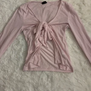 Ljusrosa långärmad topp med knyt - Supersöt ljusrosa topp med långa ärmar och v-ringning. Toppen har en snygg knyt-detalj framtill som ger en trendig look. Tillverkad i mjukt och lätt bomullsmaterial, perfekt för dig som gillar en feminin och chill stil.