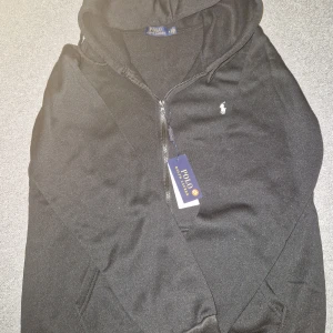 Svart hoodie från Polo Ralph Lauren - Svart hoodie från Polo Ralph Lauren med dragkedja och klassisk vit logga på bröstet. Tröjan har huva, fickor framtill och är tillverkad i mjuk bomull. Perfekt för en chill och stilren look.