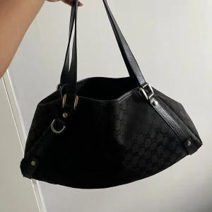 Svart axelväska från Gucci - Säljer en svart axelväska från Gucci med klassiskt GG-mönster. Väskan har detaljer i skinn och silverfärgade metalldelar. Rymlig modell med dubbla axelband och snygg form, perfekt för dig som gillar lyxiga accessoarer med ikonisk design.