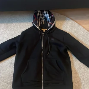 Svart Burberry hoodie med dragkedja - Svart hoodie från Burberry i storlek L med klassiskt rutigt foder i huvan. Tröjan har dragkedja framtill, snörning i huvan och broderad logga på bröstet. Perfekt för dig som vill ha en stilren och lyxig streetwear-look.