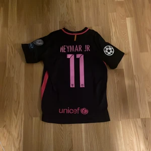 FC Barcelona Neymar Jr matchtröja - Säljer en svart FC Barcelona fotbollströja från Nike med Neymar Jr och nummer 11 på ryggen. Tröjan har rosa detaljer, klubbmärke, Champions League-märke på ärmen och Qatar Airways-tryck på bröstet. Perfekt för dig som älskar Barca och Neymar.