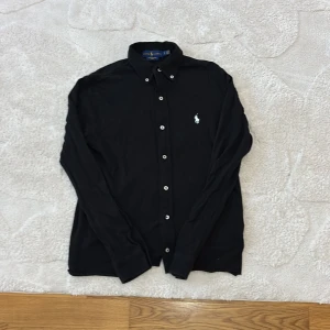 Svart skjorta från Ralph Lauren   - Klassisk svart skjorta från Ralph Lauren med vit broderad logga på bröstet. Skjortan har lång ärm, knappar framtill och är tillverkad i mjuk bomull. Perfekt för dig som gillar stilrena och tidlösa plagg.