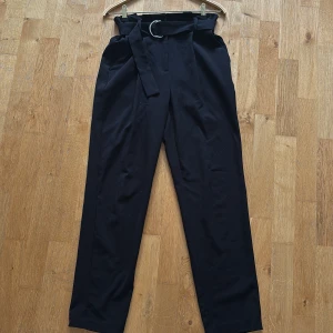 Svarta kostymbyxor med bälte - Stilrena svarta kostymbyxor med hög midja och tillhörande tygbälte med metallspänne. Byxorna har raka ben och diskreta fickor både fram och bak. Perfekta för en clean och dressad look.