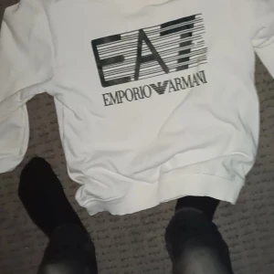 Vit sweatshirt från Emporio Armani och Boss - Två snygga vita sweatshirts, en från Emporio Armani med stor EA7-logga i svart och en från Boss med coolt tryck i svart och orange. Båda har rund hals och lång ärm, perfekt för dig som gillar stilrena märkeskläder med grafiska detaljer.