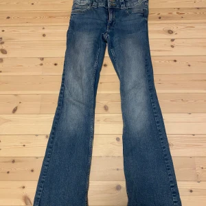 Blå bootcut jeans från H&M, low waist - Säljer ett par klassiska blå bootcut jeans från H&M knappt använda med låg midja och justerbar midja. Jeansen har snygg slitning framtill och bakfickor med knappdetalj. Perfekta för dig som gillar en avslappnad och tidlös look i storeken 170