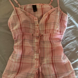 Rutig rosa topp från Vero Moda - Supersöt topp från Vero Moda i rosa och mörk rosa rutigt mönster. Klänningen har smala axelband, knappar hela vägen framtill och en snygg snörning i midjan. Perfekt för varma dagar och har en lätt, luftig känsla.( är storlek m men sitter som s)