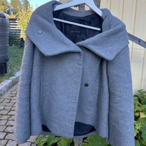 Grå ullkappa från Zara med stor krage - Snygg grå kappa från Zara med oversized krage och dold knäppning. Jackan är tillverkad i ullblandning och har raka ärmar samt två diskreta fickor framtill. Liten i storleken