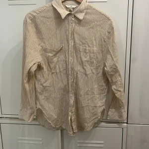 Beige och vit randig skjorta H&M - Säljer en beige och vit randig skjorta från H&M i 100% bomull. Skjortan har klassisk krage, knäppning framtill, två bröstfickor och långa ärmar. Perfekt för en chill och avslappnad stil.
