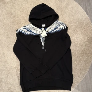 Svart hoodie Marcelo Burlon med vingar - Svart hoodie från Marcelo Burlon County of Milan med ikoniskt vingtryck i vitt och blått över axlar och bröst. Tröjan har huva med snörning och känguruficka framtill. Tillverkad i mjuk bomull för en skön och avslappnad stil. Helt ny och inte använd en ända gång säljer på grund av stor i storleken.