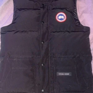 Svart dunväst från Canada Goose - Säljer en svart dunväst från Canada Goose med klassisk logga på bröstet. Västen har tryckknappar framtill, två stora fickor och är perfekt för lager-på-lager. Tidlös design och riktigt varm, passar dig som vill ha något både snyggt och funktionellt.