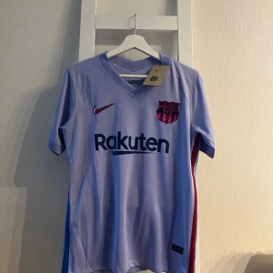 FC Barcelona bortatröja Nike S - Säljer en FC Barcelona bortatröja från Nike i ljuslila med klubbmärke, Rakuten-tryck och La Liga-patch på ärmen. Tröjan är i funktionsmaterial (Dri-FIT) och har korta ärmar samt röda och blå detaljer på sidorna.