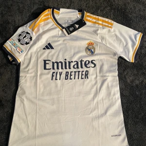 Real Madrid Mbappé #10 vit matchtröja - Real Madrid fotbollströja med Mbappé och nummer 10 på ryggen. Vit med guld- och mörkblå detaljer, korta ärmar och broderade klubbmärke samt Champions League-patch. Tillverkad i lätt polyester med Aeroready-teknologi. Snygga Adidas-ränder på axlarna.