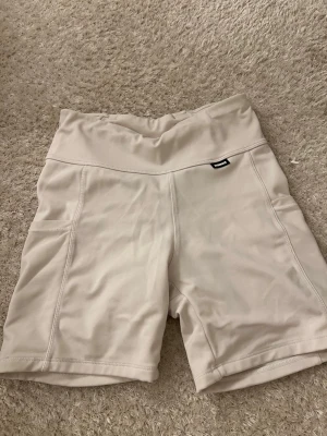 Beige träningsshorts från Stronger - Säljer ett par ljusbeige träningsshorts från Stronger med hög midja och praktiska sidofickor. Shortsen är tillverkade i ett stretchigt syntetmaterial som sitter skönt och följer kroppen. Perfekta för gymmet eller löprundan.