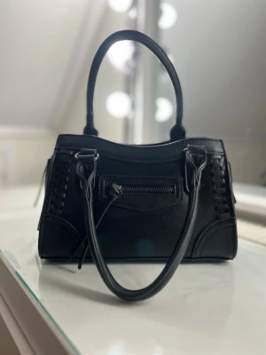 Svart handväska med dragkedja och snygga detaljer från Gina Tricot💕 - Säljer en praktisk och snygg svart handväska från Gina Tricot. Den är använd ungefär 5/6 gånger men är som ny. Den är även nydligen tvättad på utsidan🫶🏻OBS! Band tillkommer tyvärr inte då jag tappat bort det.