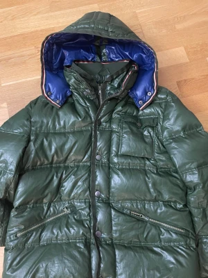 Grön glansig pufferjacka - Säljer en mörkgrön pufferjacka med blank yta och blått foder i huvan. Jackan har dragkedja och knappar framtill, flera fickor med dragkedja och knapp, samt snygga randiga detaljer längs huvkanten. Perfekt för kalla dagar och riktigt skön att bära. Passformen är som fjällräven jackan, alltså lite längre i modellen!