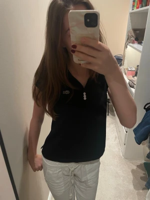 Svart pikéblus från Lacoste - Svart pikéblus från Lacoste med klassisk krage, tre vita knappar framtill och det ikoniska krokodilmärket på bröstet. Kortärmad och i mjuk bomull, perfekt för en clean och sportig look.