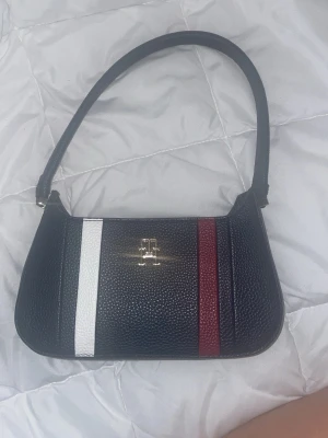 Blå vintage axelväska från Tommy Hilfiger - Snygg svart axelväska från Tommy Hilfiger i skinn med struktur. Väskan har ett stilrent randigt mönster med vita och vinröda detaljer på framsidan samt ett metallfärgat TH-emblem. Perfekt storlek för det viktigaste och har en klassisk form.