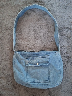 Blå axelväska i denim - Säljer en blå axelväska i denim med en bred axelrem och praktisk dragkedja upptill. Väskan har en stor ficka framtill med knappstängning och är perfekt för att bära med sig det viktigaste. Enkel och cool design som passar till jeans eller streetwear.