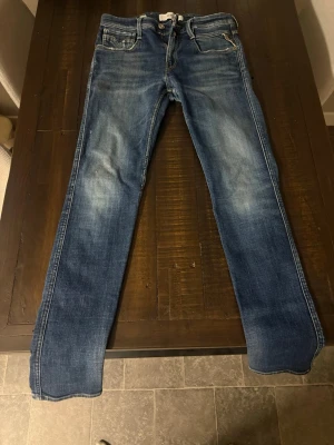 Replay Anbass slim fit jeans blå - Säljer ett par Replay Anbass slim fit jeans i klassisk blå tvätt med snygga slitningar och kontrastsömmar. Modellen har fem fickor, dragkedja och knapp i midjan samt detaljerade bakfickor. Perfekt passform för dig som gillar smalare jeans.