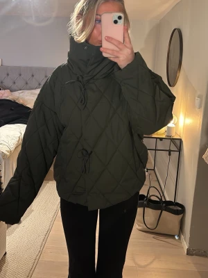 Mörkgrön quiltad pufferjacka - Säljer en mörkgrön pufferjacka med quiltat mönster och hög krage. Jackan har oversized passform, långa ärmar och snygga snörningsdetaljer framtill. Perfekt för kyliga dagar och riktigt trendig look.