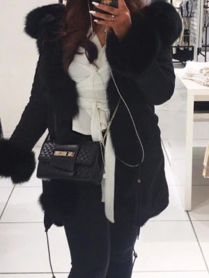 Svart pälsparkas  - Säljer en svart parkajacka från moomookong med fluffiga pälsdetaljer på huva, krage och ärmslut. Jackan har snörning nedtill och en loose passform som ger en cool vibe. Perfekt för kalla dagar när du vill vara både varm och snygg. Köptes för 3100kr.