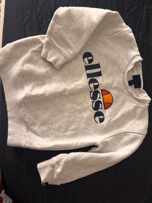Vit sweatshirt från Ellesse - Säljer en vit sweatshirt från Ellesse med stort tryck på bröstet i marinblått, rött och orange. Tröjan har rund hals, ribbade muddar vid ärmslut och nederkant samt långärmad passform. Perfekt för dig som gillar sportiga och klassiska märken.