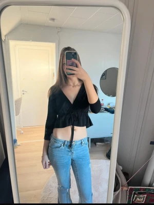 Svart Gina topp - Säljer en svart omlottopp med v-ringning och snörning i midjan. Toppen har långa ärmar och en volangkant nedtill som ger en snygg siluett. U mycket bra skick och inga defekter! I storlek Xs