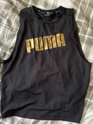 Svart linnetopp från Puma med snörning - Titta in på min sida för liknande. Säljer en svart linnetopp från Puma i storlek L. Toppen har ett stort guldigt Puma-tryck framtill och snörningsdetalj med knyt baktill. Materialet är mjukt och känns som bomull, perfekt för träning. Ärmlös och loose fit för skön rörelsefrihet.
