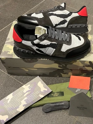 Valentino Garavani Rockrunner sneakers - Säljer ett par Valentino Garavani Rockrunner sneakers med svart, vit och grått camouflage-mönster, samt röd detalj på hälen. Skorna har snörning, platt sula med nitar och är tillverkade i en mix av skinn, mocka och textil. Kommer med originalkartong, extra skosnören och dustbag.