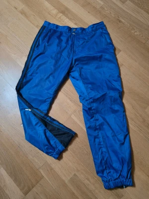 Blå täckbyxor med svarta ränder - Säljer ett par blå täckbyxor från Adidas med svarta ränder längs sidorna. Byxorna har resår i midjan och benslut, samt dragkedjor vid anklarna för extra ventilation. Materialet är syntet och passar perfekt för utomhusaktiviteter.