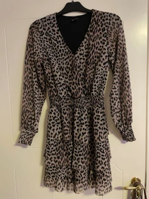 Leopardmönstrad kortklänning Gina Tricot - Snygg kortklänning från Gina Tricot i leopardmönster med beige och svarta toner. Klänningen har v-ringad omlottfront, långärmade ärmar med smock vid handlederna och markerad midja med smock. Materialet är lätt och luftigt, perfekt för att sticka ut.