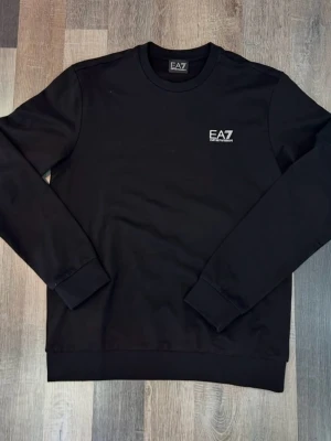 Svart sweatshirt från EA7 Emporio Armani strl M - Mycket gott skick.  Svart sweatshirt från EA7 Emporio Armani med vit logotyp på bröstet och vita ränder i nacken på ryggen. Tröjan har rund hals, långa ärmar och ribbade muddar vid ärmslut och nederkant. Perfekt för en stilren och sportig look.