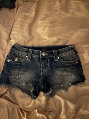 True Religion Joey Cut-Off jeansshorts - Säljer ett par blå denimshorts från True Religion, modell Joey Cut-Off. De är lågmidjade med slitna detaljer och fransad kant. Klassiska femficksmodellen med snygga silverknappar och broderade bakfickor. Perfekta för sommaren och riktigt sköna att ha på sig.