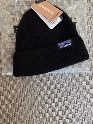 Svart ribbad mössa från Patagonia - Säljer en svart ribbstickad mössa från Patagonia med uppvikt kant och klassisk logga framtill. Perfekt för kalla dagar och enkel att matcha med olika outfits.