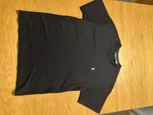 Svart t-shirt från Polo Ralph Lauren - Svart t-shirt från Polo Ralph Lauren i mjuk bomull. Klassisk rund hals och korta ärmar. På bröstet finns den ikoniska broderade polospelaren i vitt. Perfekt basic med stilren design och skön passform.